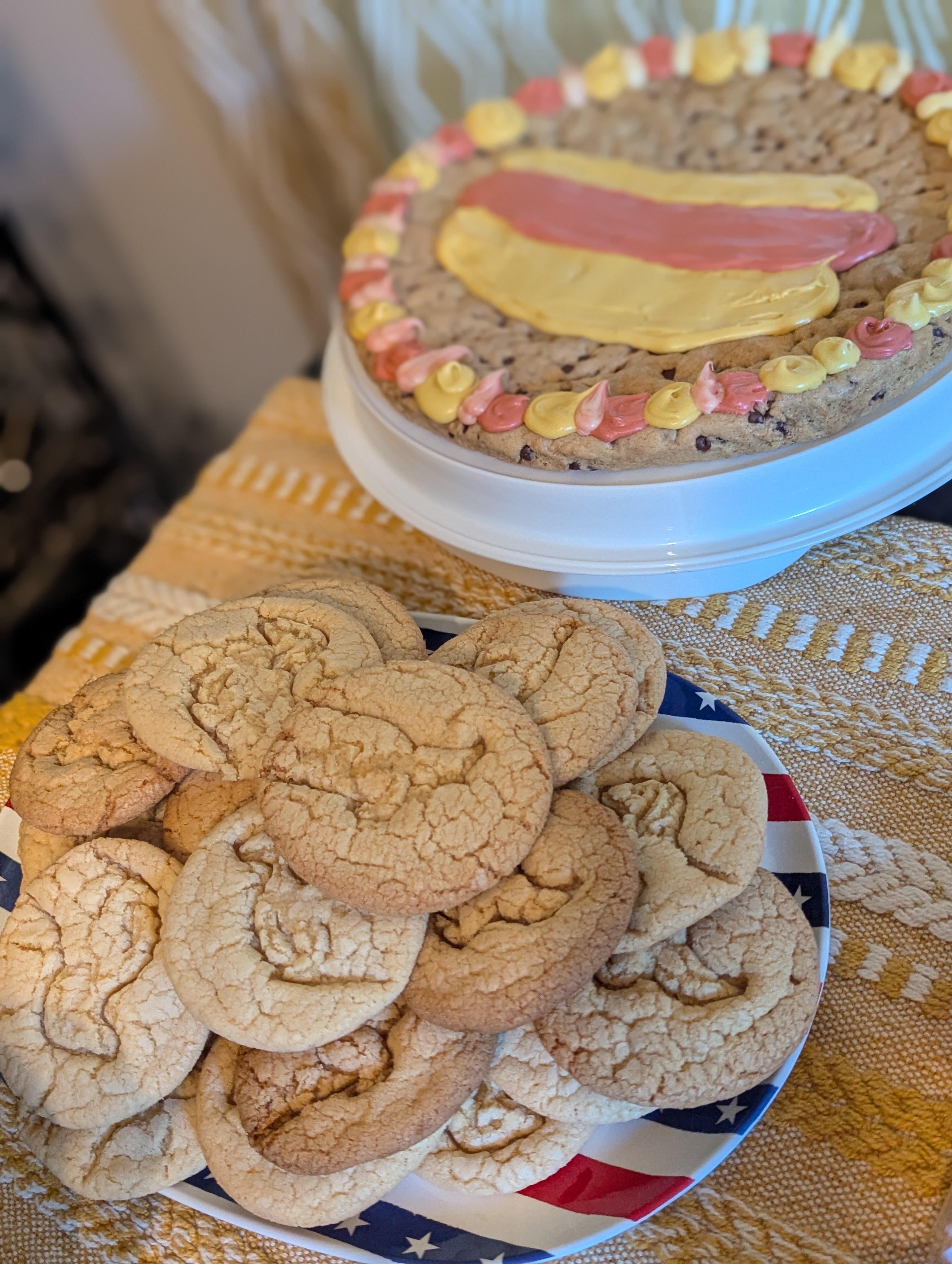 A plate of Paula Deen sugar cookies with the shape of a dachshund stamped into them. Dachshunds are long, so half of the cookies have the shape of a dog's head and little legs and the other half of the batch has wiggle-butts. Also pictured: a supermarket cookie cake decorated to resemble the hotdog emoji.